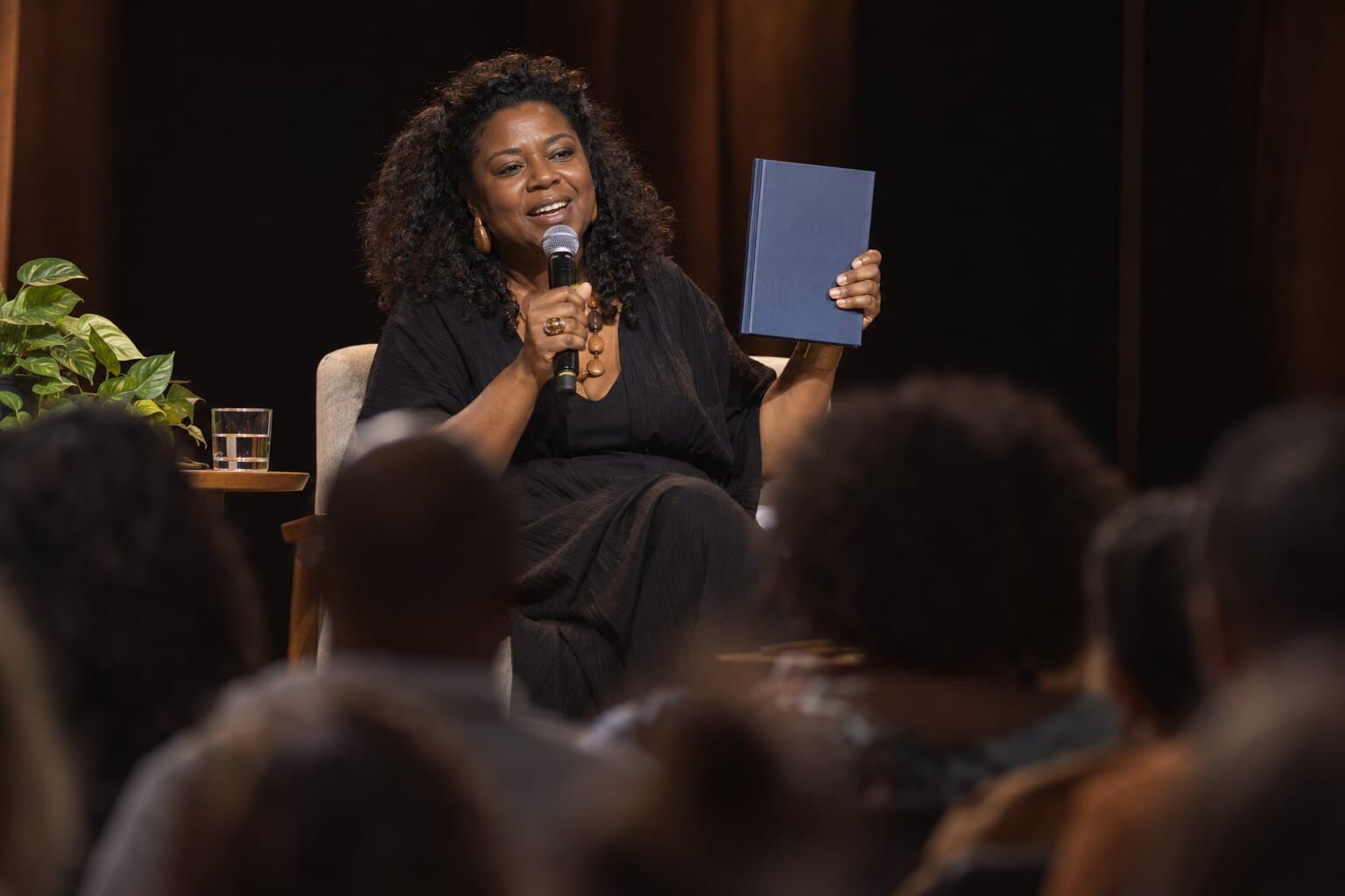 Uma autora fala num palco, sorrindo, com o próprio livro erguido na mão diante da plateia.