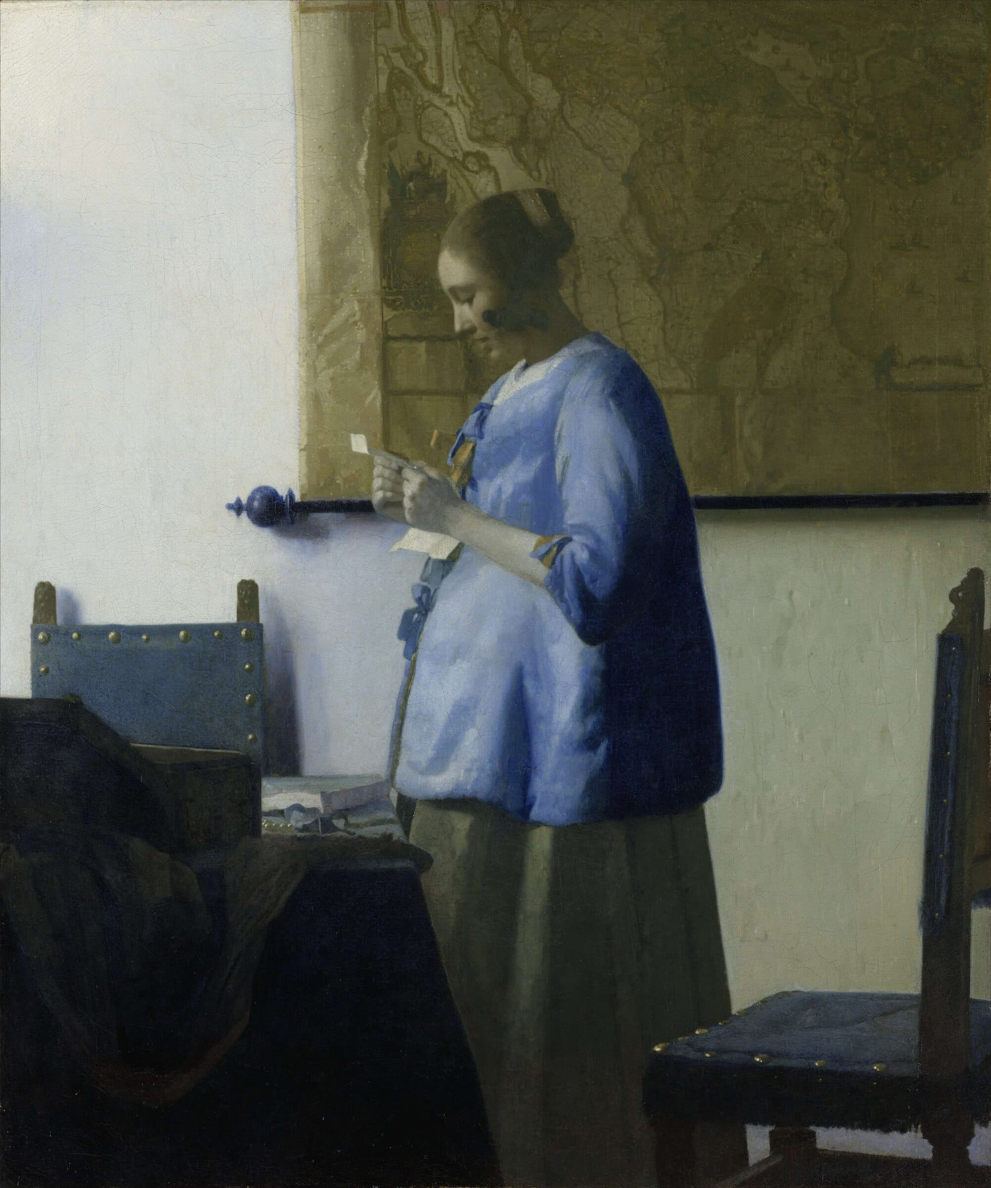 Mulher de casaco azul lê uma carta de pé, diante de um mapa na parede.