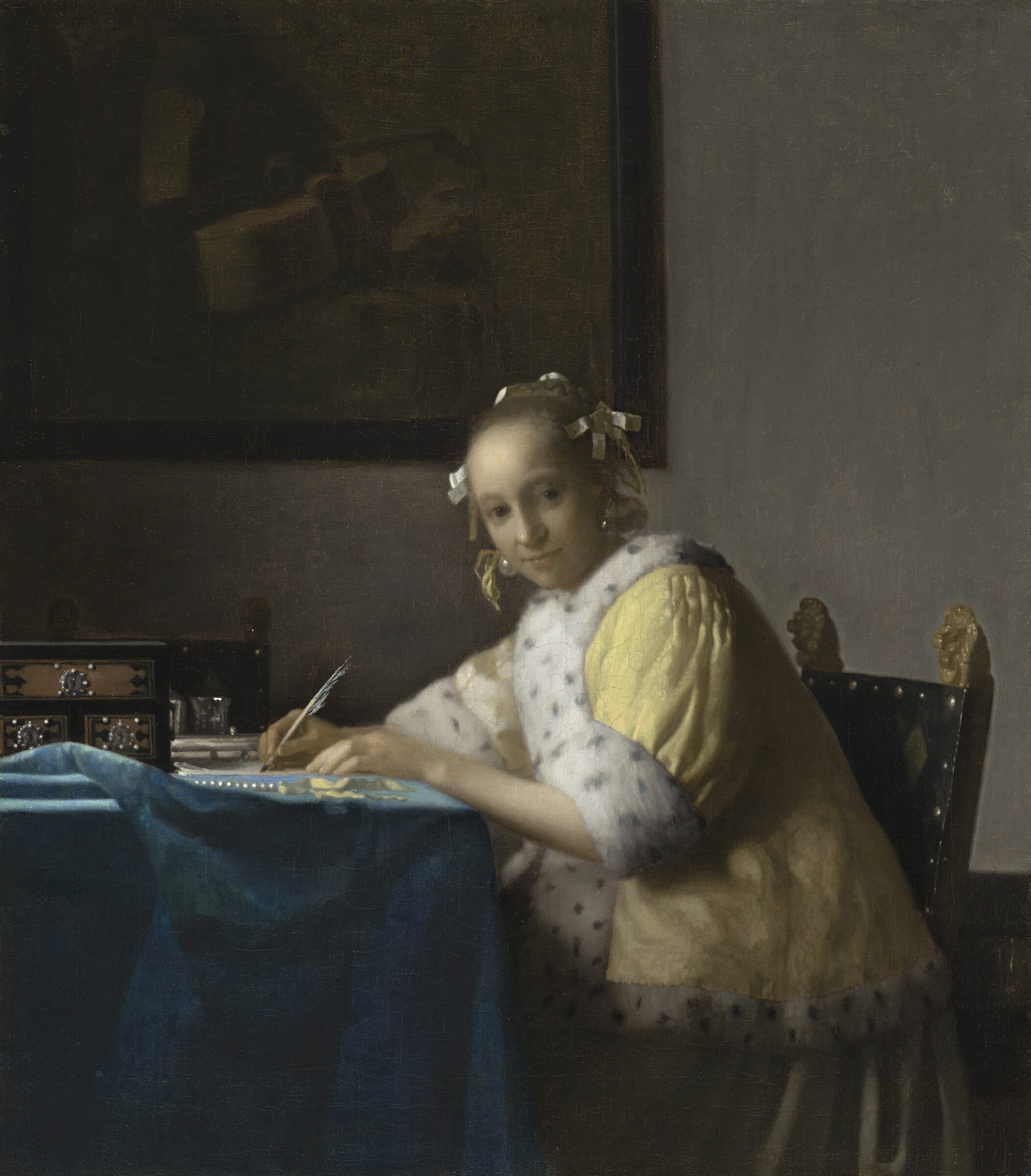 Mulher de casaco amarelo escrevendo uma carta à mesa ergue os olhos e encara quem a observa.
